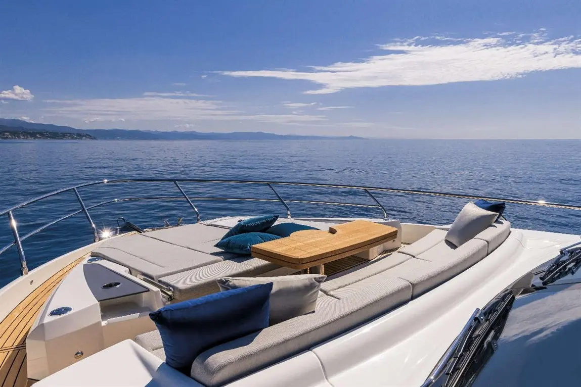 Luxurious yacht deck with cushions and a wooden table, overlooking calm sea and distant mountains under a clear blue sky.